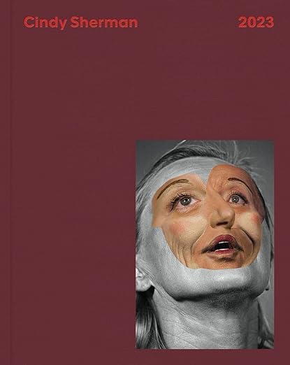 A dark red book cover with a photograph of a woman with a half-painted face. The title reads "Cindy Sherman: 2023."
