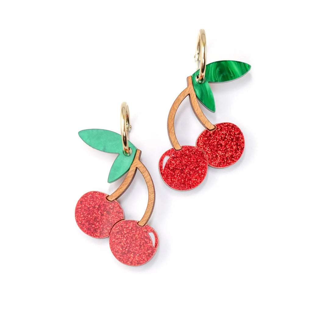 A pair of cherry earrings with red glitter. They are hanging on golden hoops.
