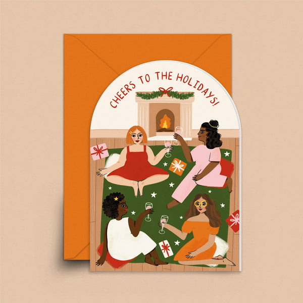 An arch-shaped greeting card with an orange envelope. The card features four diverse women sipping a drink in front of the fireplace during the holidays under the words "Cheers to the holidays."