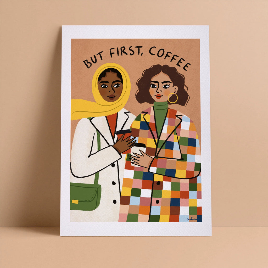 A print with a white border around the illustration. The illustration features a woman with a hijab and a dark skin-tone next to a woman with short, curly, brown hair and a medium-skin tone. They are both wearing trench coats and sipping coffee. They are before a beige background with the words "But First, Coffee."