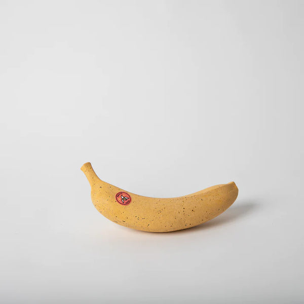A white background with a yellow banana before it. The banana is freckled with brown dots. There is also a grocery store banana sticker on it with the letters "P" and "C."