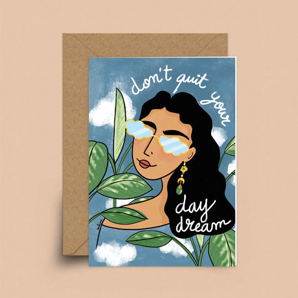 A blue greeting card with a brown envelope. The card depicts a blue sky in the background. Before it is a woman with a medium skin-tone and wavy, dark hair wearing cloud sunglasses. She is sitting within a few leaves with the words "Don't Quit Your Day Dream" above her head. 
