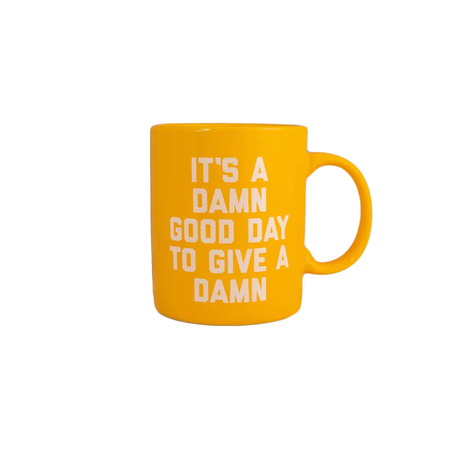 A white background with a yellow mug before it. The mug is yellow with the words "It's A Damn Good Day To Give A Damn" written on it in white letters.