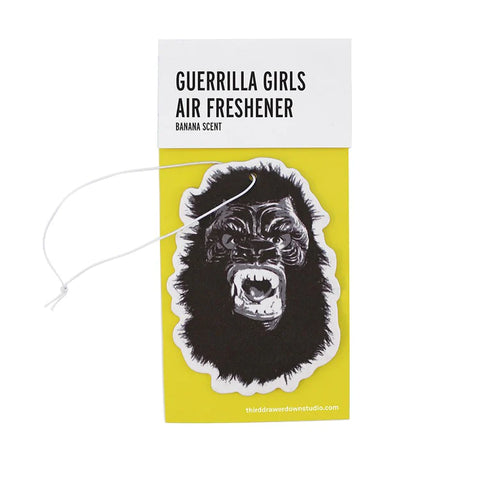 A white background with a yellow package in front of it. The yellow package holds an air freshener in the shape of a gorilla with its mouth open. On the top of the package is a white strip of packaging that holds everything together. On the white strip are the words "Guerrilla Girls Air Freshener" and the words "Banana Scent" in smaller letters. 