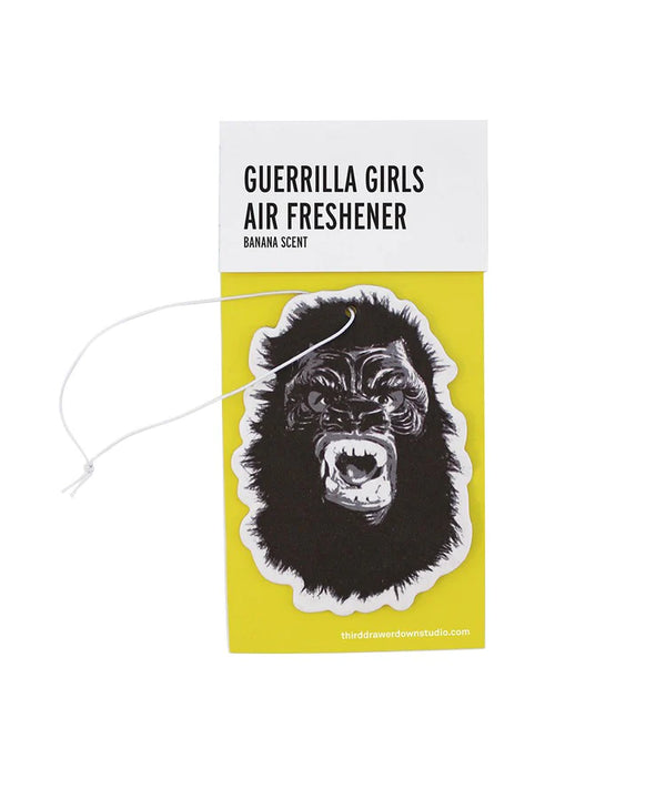 A white background with a yellow package in front of it. The yellow package holds an air freshener in the shape of a gorilla with its mouth open. On the top of the package is a white strip of packaging that holds everything together. On the white strip are the words "Guerrilla Girls Air Freshener" and the words "Banana Scent" in smaller letters. 