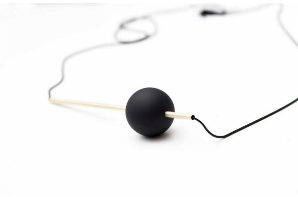 A white background with a necklace in front of it. The necklace is a black, circular bead with a gold bar running through it. The bar and orb are held together by a black chain. 