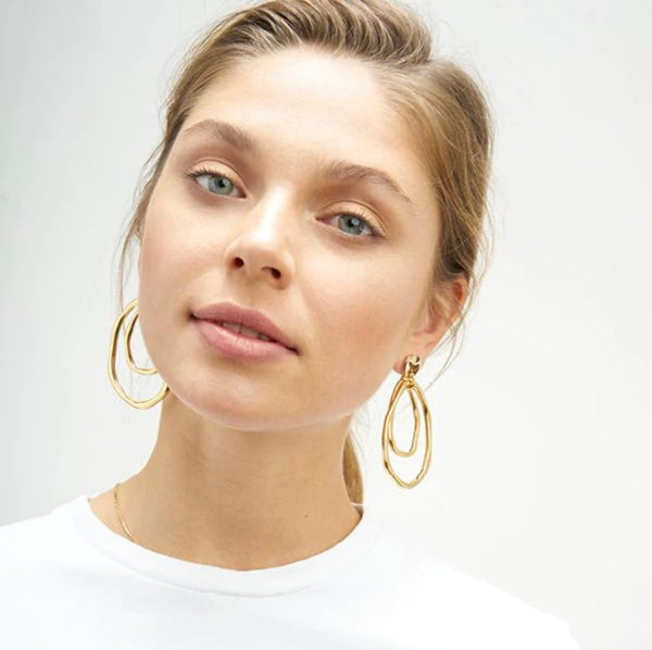 A woman with a light skin tone and blonde hair is wearing golden earrings. They are stud earrings with two hanging irregular circle pieces, and one bigger than the other.