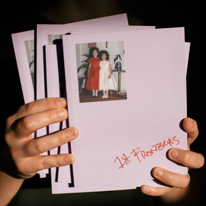 A book with a lavender cover. The book features a picture in the top left of two young girls with curly, brown hair. One girl is in a red dress and the other is in a white dress. Handwritten on the bottom right corner is the title "First Fronteras."
