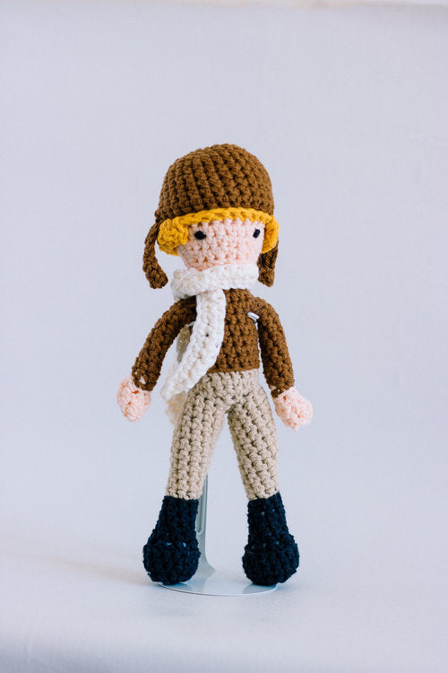 A crochet doll of a person with blonde hair and a pilot's uniform in brown.