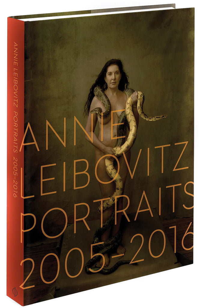 A charcoal book cover with a red spine. Featured in the foreground of the backdrop is Marina Abramovic nude and holding two snakes. The title, "Annie Leibovitz Portraits 2005-2016" is written in large, orange letters. 