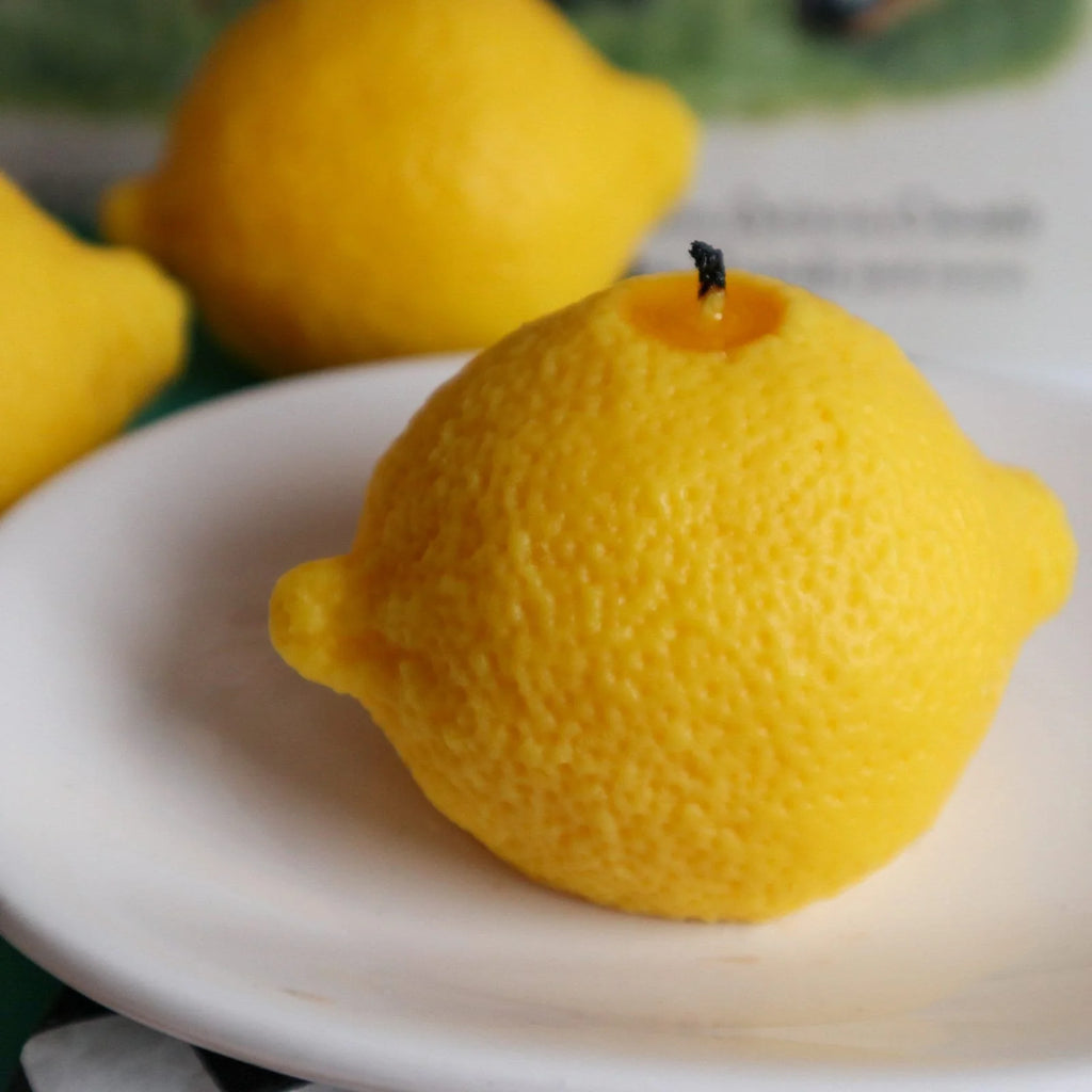 A small white plate with lemons that are blurred in the background. On the plate is a candle with the appearance of a lemon, and a slightly burnt wick. 