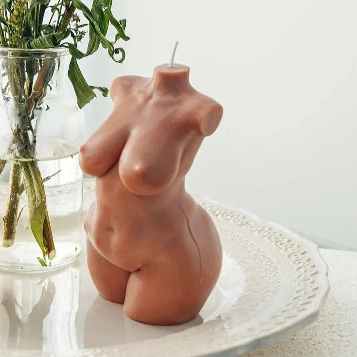 A white background that features a plant in a clear vase filled with water. Before it is a candle in the shape of a nude woman with a medium skin-tone. The candle sits on a white plate. 