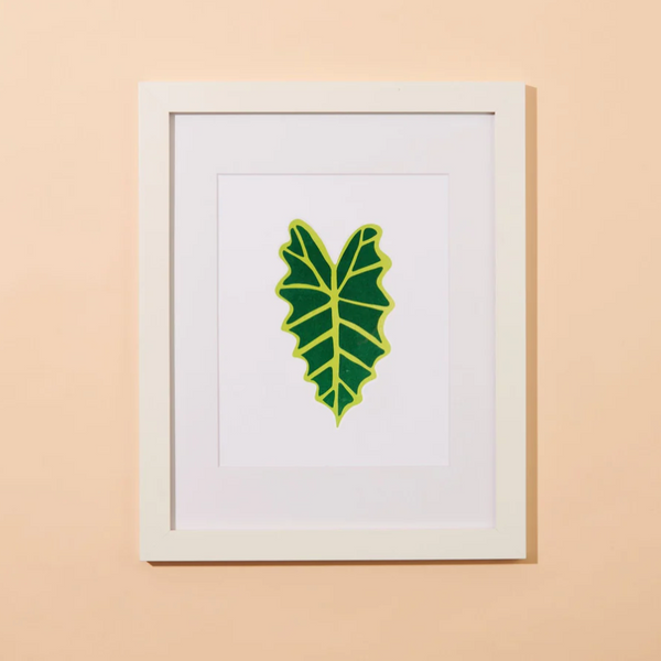A peachy background with a framed print before it. The print features a singular alocasia leaf. 
