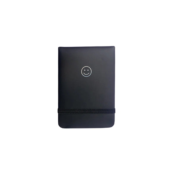 A white background with a black notepad in front of it. The notepad features a silver smiley face and an elastic band around it. 