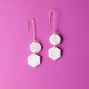 A set of two white, geometric earrings before a pink background. They are attached to a rose-gold hook.