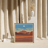 A print of a desert landscape with a mountain range in the far back. The print is framed and standing before curtains.
