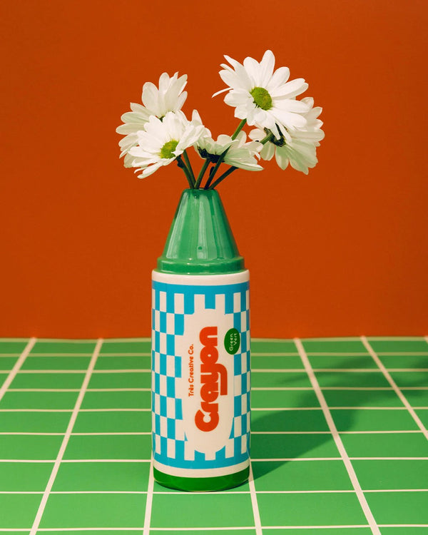 A hand-painted vase resembling a crayon in green with a blue checkered pattern before a red background, holding white flowers.