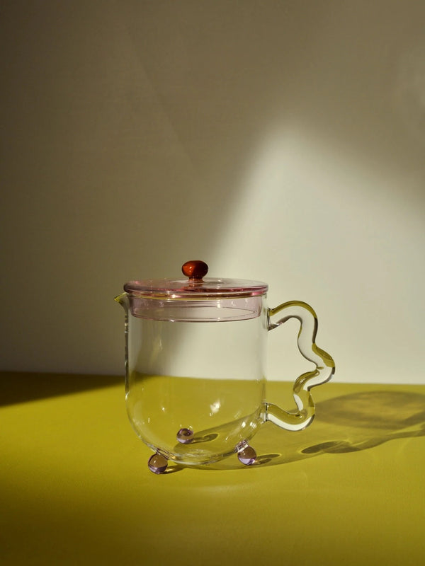 A small teapot with a pink tinted lid and a green squiggly handle. The pot is standing on a green surface.