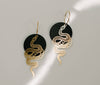 A snake earring. The snake is golden and attached to a black circle onto a golden hoop.