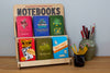 A wooden book stand with several notebooks with colorful covers. Next to it are two vessels with pencils in them.