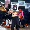 A woman is holding a mug and a pink clutch that reads: "The Advantages of being a woman artist." She is wearing a bag over her head with a print of a gorilla head. Behind her is a life-size photograph of women in gorilla masks plastering walls in posters.