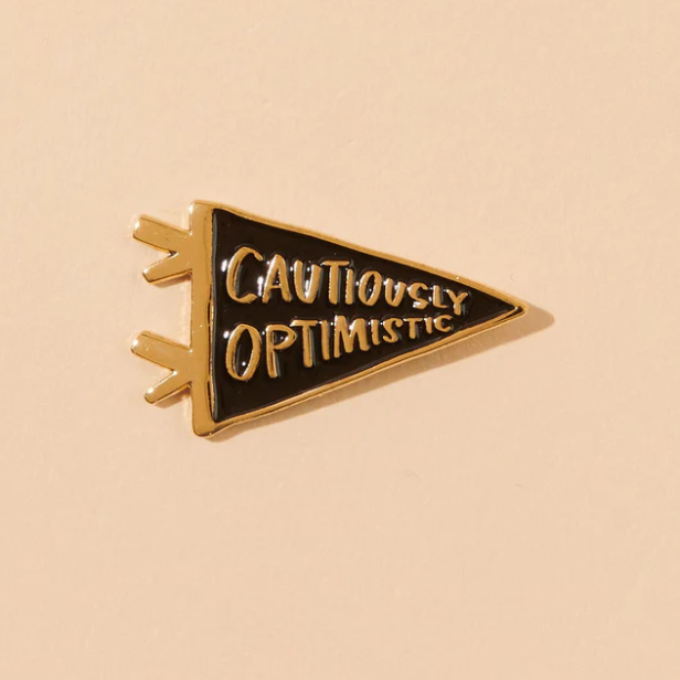 A peachy background with a black pin before it. The pin is in the shape of a flag with the words "Cautiously Optimistic" written in gold. 