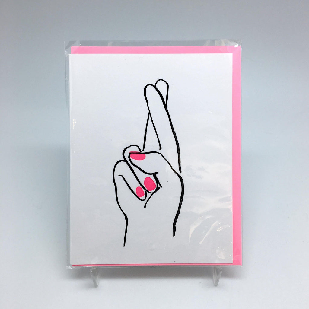 A white background with a white card before it. The card features a hand with crossed fingers and neon pink nails. 