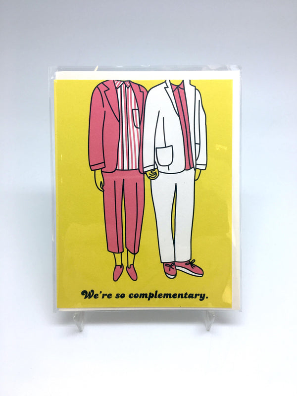 A yellow greeting card with an illustration of two people in a white and a pink suit holding hands. The text reads "We're so complementary."