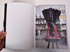 Look inside a  book with a photograph of the inside of a ceramic studio. A black sculpture is on its head on a table.