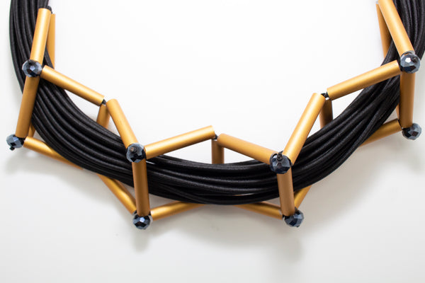 A black and gold necklace made from cords and golden brushed aluminum parts creating geometric shapes around the necklace.