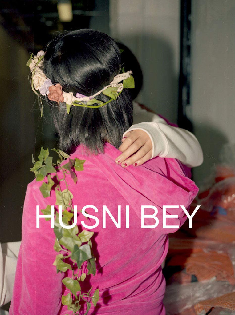 Book cover featuring a photograph of a woman from behind wearing a floral crown and a pink hoodie, holding a child. The title reads: "Husni Bey."