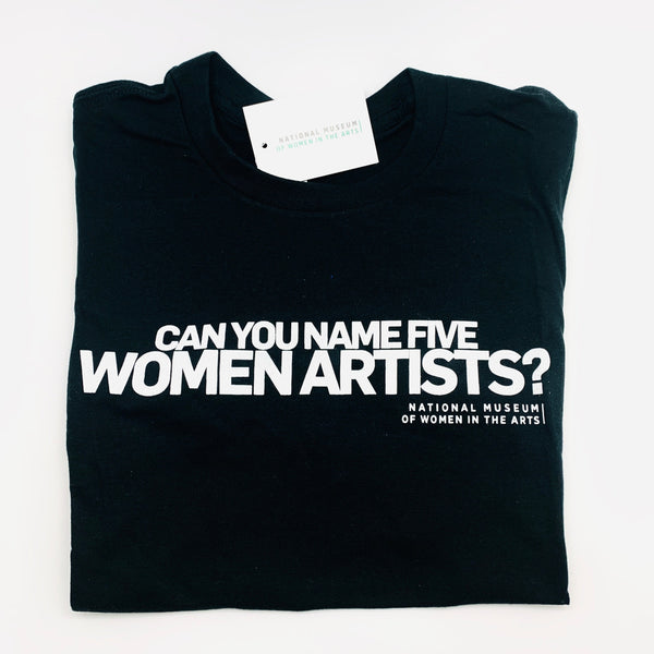A folded black t-shirt is lying on a white background. On the t-shirt, text in large white capital letters reads: "Can you name five women artists?"