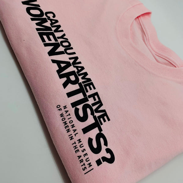 A folded pink t-shirt is lying on a white background. On the t-shirt, text in black capital letters reads: "Can you name five women artists?"