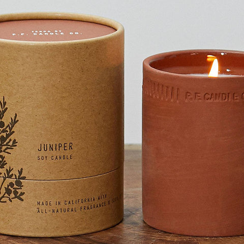 A round paper box with the text "Juniper: Soy Candle" next to a candle in a red clay cup, burning.