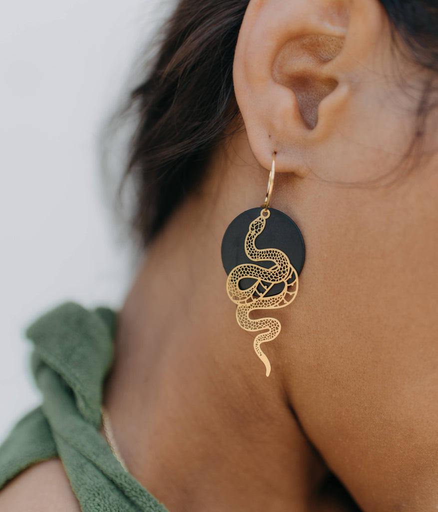 A woman with a medium skin tone wearing a snake earring. The snake is golden and attached to a black circle onto a golden hoop.