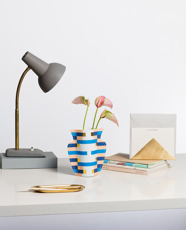 A paper flower vase with an abstract pattern in blue and peach standing on a table next to a desk lamp and books.