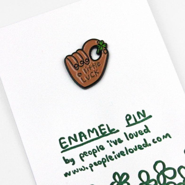 A little enamel pin on a white card. The pin depicts a dark-skinned hand holding a clove. On the hand, it says: "A little luck."